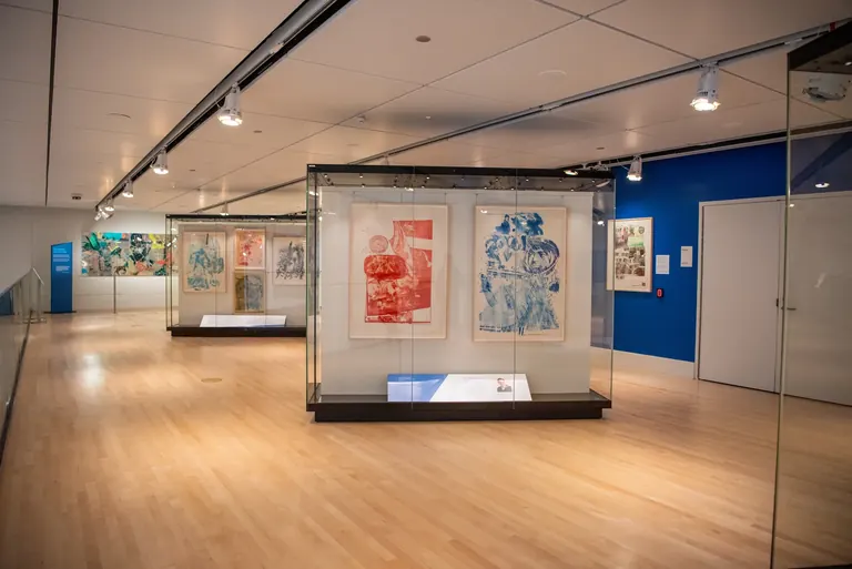 Two display cases inside a museum gallery with rich blue walls; the case in the foreground holds two Rauschenberg works and the one behind it holds four
