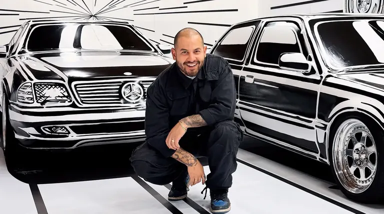 L’artiste Joshua Vides accroupi entre deux voitures peintes à la main dans son style de contours noirs et blancs