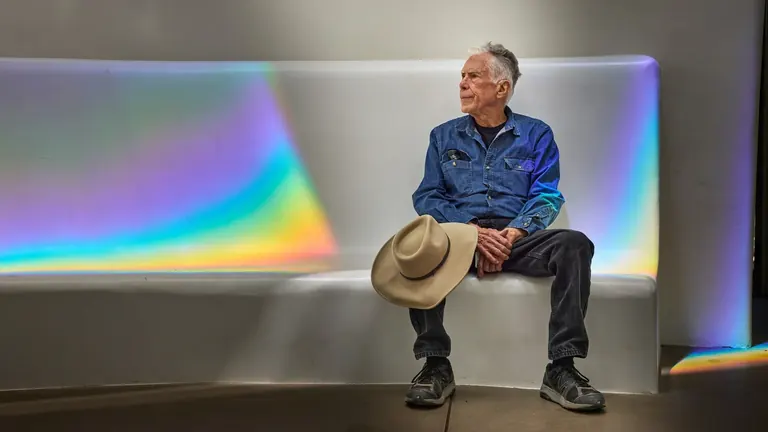 Un homme est assis sur un banc blanc tandis que les arcs-en-ciel lumineux de l’installation prismatique de Charles Ross se déploient autour de lui