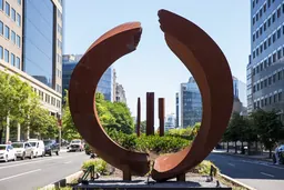 A circular, rust-colored sculpture and three rust-colored columns in the median of a city street.