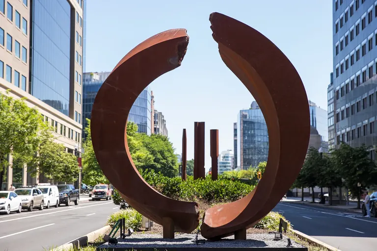 A circular, rust-colored sculpture and three rust-colored columns in the median of a city street.