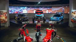 Voitures de course tout-terrain sur une plateforme surélevée, derrière trois motos de motocross rouges, devant un large écran vidéo montrant une course sur terre