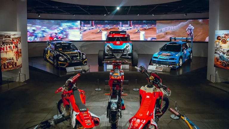 Voitures de course tout-terrain sur une plateforme surélevée, derrière trois motos de motocross rouges, devant un large écran vidéo montrant une course sur terre