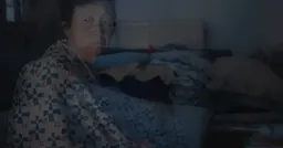 A translucent film still of an older person wrapped in a blue, white and red patterned blanket, in a dimly lit room with a radiator and piled blankets