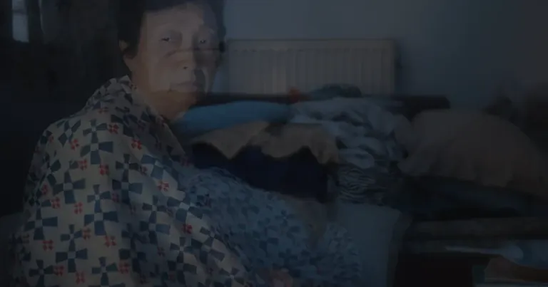 A translucent film still of an older person wrapped in a blue, white and red patterned blanket, in a dimly lit room with a radiator and piled blankets