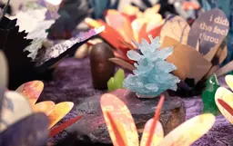 A close-up of a small, light blue crystal formation displayed on a rock, surrounded by colorful, paper-like flower shapes and decorations on a purple textured surface.