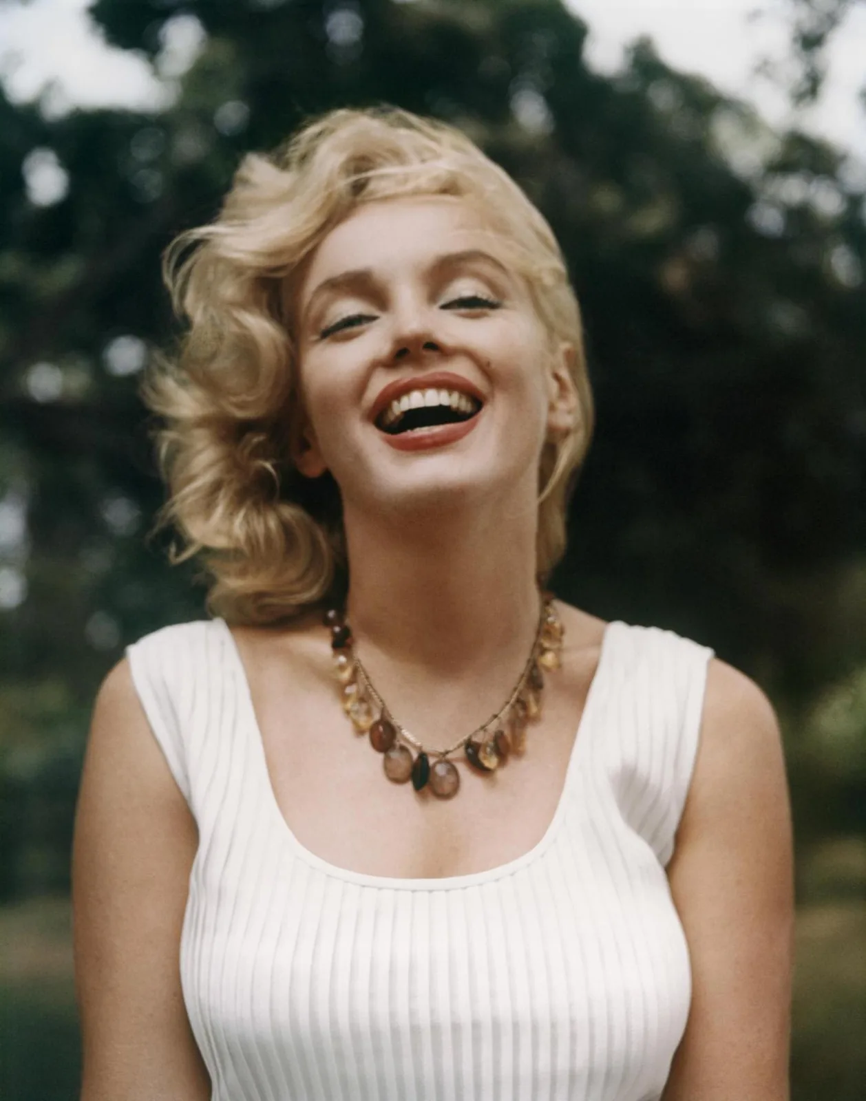 Colour photograph of Marilyn Monroe from the waist up, short blonde hair blowing in the wind, parted red lips, wearing a white ribbed vest and beaded necklace.