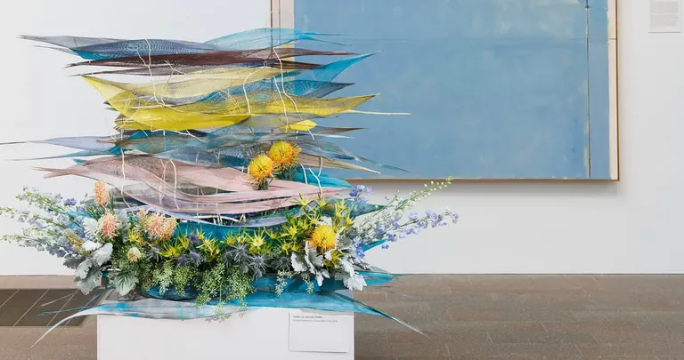 Une composition florale posée sur une sculpture tressée bleue et jaune, devant une peinture abstraite