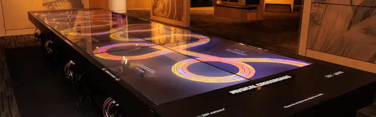 Taula tàctil interactiva de Musical Crossroads que mostra línies del temps dels gèneres musicals en bucle, amb auriculars al llarg de la vora