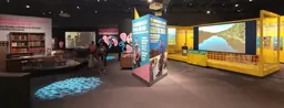 Expositions du musée consacrées à deux scientifiques. À gauche, un bureau et des étagères de livres, avec des panneaux multimédias. À droite, un grand écran vidéo et un canoë.