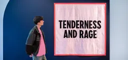 Una visitant a l’entrada de l’exposició mira un gran cobrellit rosa clar amb la vora vermella intensa, on hi ha cosides en majúscules les paraules «Tenderness and Rage»