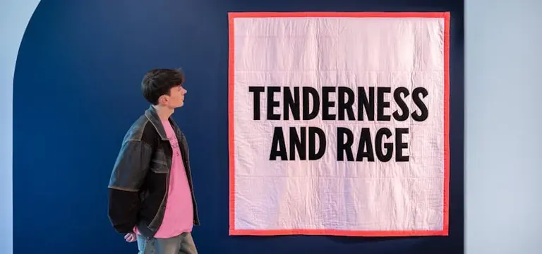 Una visitant a l’entrada de l’exposició mira un gran cobrellit rosa clar amb la vora vermella intensa, on hi ha cosides en majúscules les paraules «Tenderness and Rage»