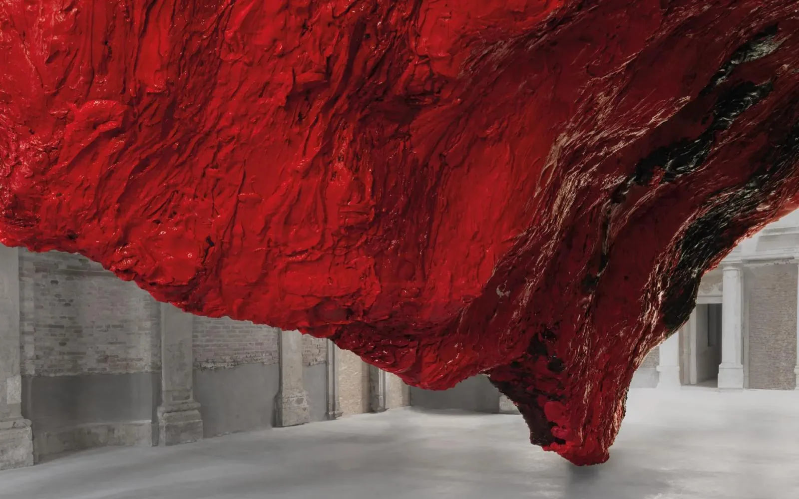Large red textured sculpture by Anish Kapoor hanging from ceiling to floor in a columned space