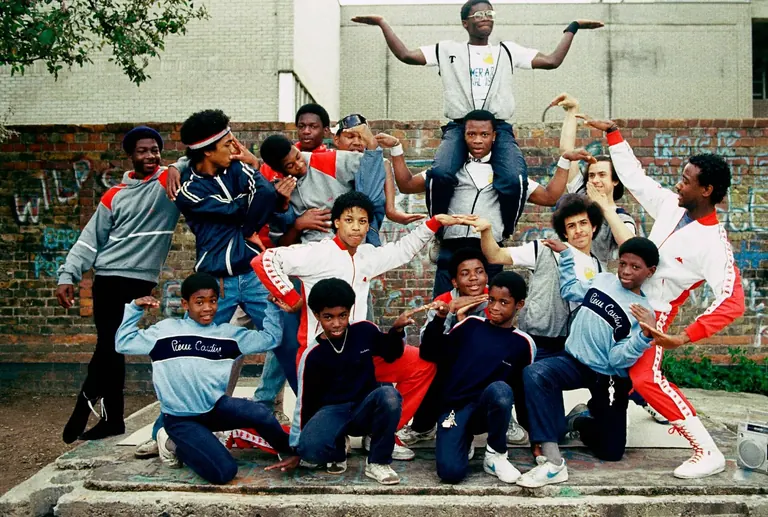 A group of young breakdancers in tracksuits posing in front of a graffitied brick wall, London, 1983