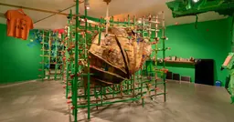 A large, textured, brown cardboard sculpture encased within a green wooden grid structure, in a room with bright green walls and an orange t-shirt hanging from the ceiling
