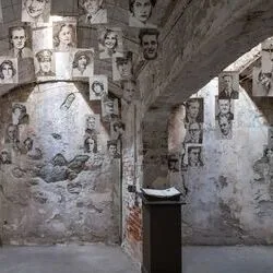Cindy Stockton Moore, Other Absences, in einer Gefängniszelle mit abblätternder Farbe und freiliegendem Mauerwerk; Porträts an Wänden und Decke; ein Buch auf einem Sockel.