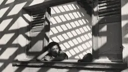 Photographie en noir et blanc d’une femme penchée à une fenêtre, dans l’ombre d’un treillis quadrillé