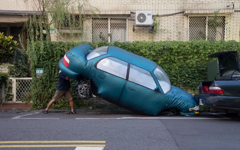 Un hombre parece levantar la parte trasera de una gran réplica inflable de un coche, de color verde azulado, encajada en una plaza de aparcamiento junto a la acera, delante de un edificio residencial