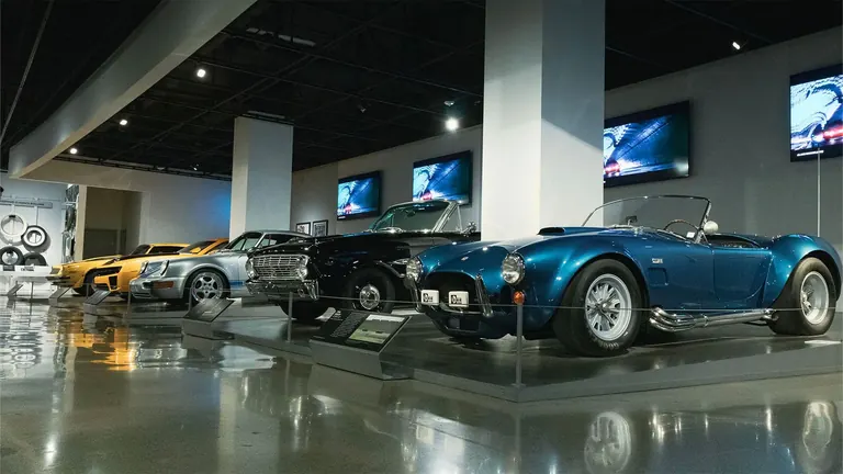 Rangée de voitures de cinéma, en tête de laquelle se trouve un roadster Shelby Cobra bleu ; au-dessus, des écrans diffusent des extraits de films
