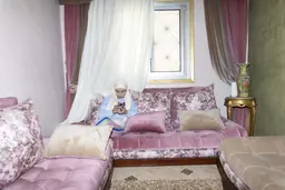 Une femme assise sur un canapé rose à motifs floraux regarde son téléphone, dans la série In Her Own Way de Yomna El Beyaly