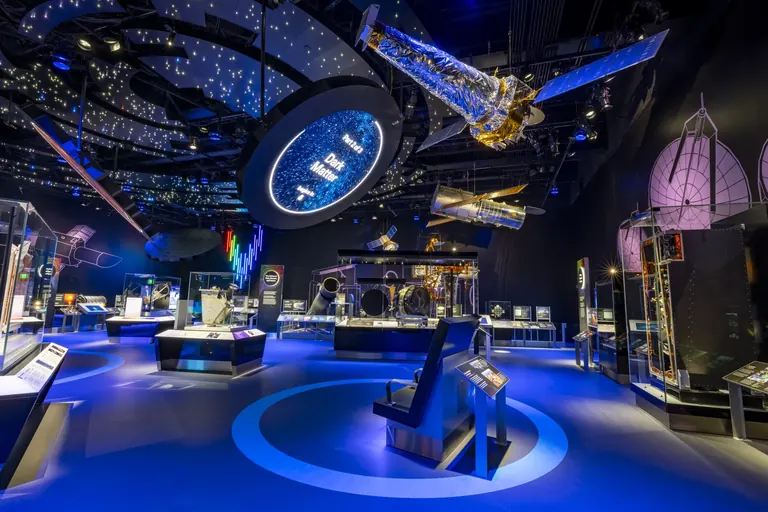 A gallery lit in purples and blues with a space telescope model suspended from a ceiling dotted with star-like lights, and object cases throughout