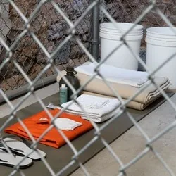 Installation „GTMO“ von William Cromar in einem eingezäunten, beengten Raum mit Eimern, gefalteten Decken, einem orangefarbenen Kleidungsstück, Flip-Flops und Toilettenartikeln.