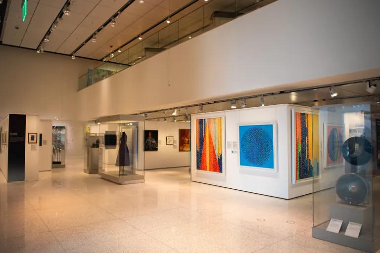 A gallery hung with paintings; two glass sculptures sit in a display case in the foreground and a purple dress is displayed in a case behind