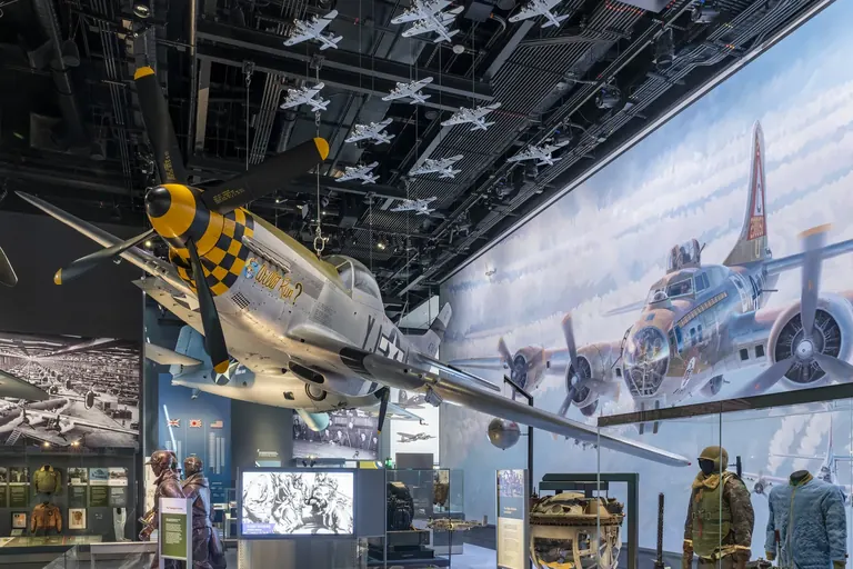 A silver fighter aircraft with a black and yellow checkered nose suspended at an angle from the ceiling, against a wall-spanning mural of a bomber in flight, with uniforms displayed below