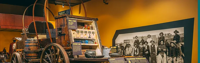 修復後的牧牛篷車，旁邊放有標示清楚的補給箱；它位於 Cowboy Gallery，旁邊是一幅大型歷史照片，攝有一群牛仔