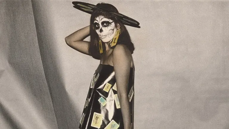 Une personne maquillée en squelette et vêtue d’une robe porte des glaïeuls et pose devant un fond drapé, dans Lotería de Muertos de Ricardo Valverde