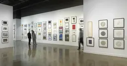Installation view with framed artworks hung salon-style on the walls and three people looking at the art