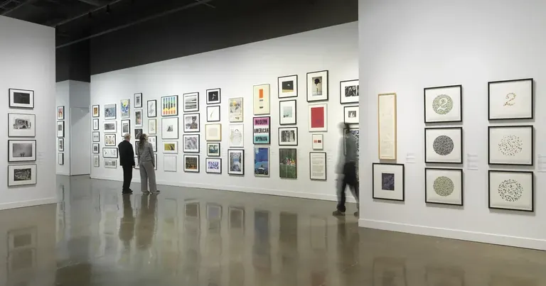 Installation view with framed artworks hung salon-style on the walls and three people looking at the art
