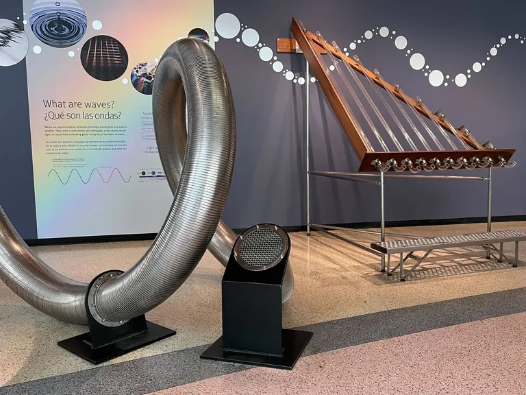 Un Echo Tube metàl·lic enrotllat i l’exposició Pipes of Pan davant d’un plafó mural amb la pregunta «What are waves?»