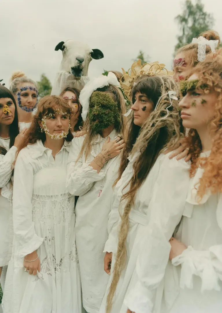 Fotografia en color d’un grup de dones joves a l’aire lliure, amb vestits blancs llargs i la cara decorada amb flors i molsa; al darrere, algú sosté enlaire un cap d’ovella de joguina.