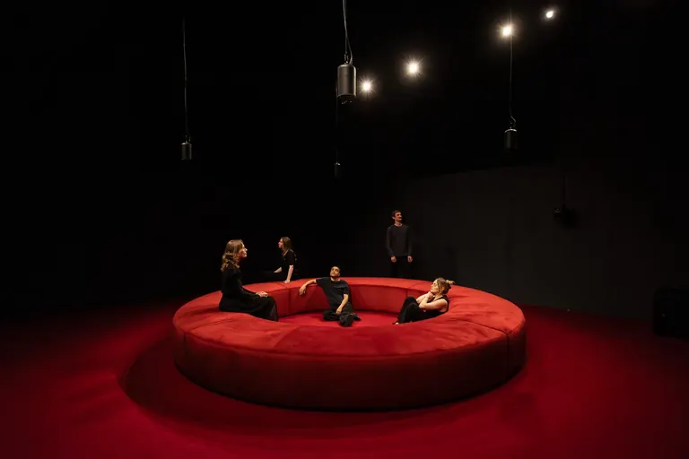 Des visiteurs assis sur un banc circulaire rouge dans la salle d’écoute spatiale 4DSOUND plongée dans l’obscurité, au Nxt Museum, Amsterdam