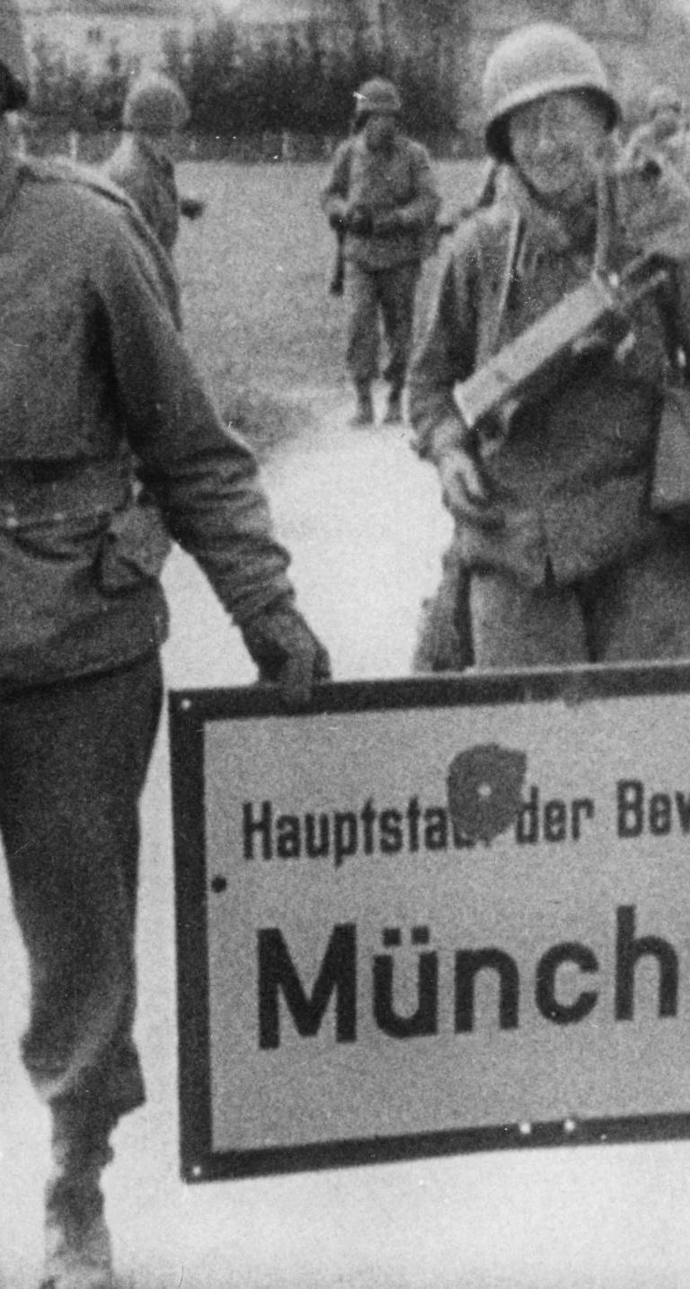 Three armed American soldiers carrying a place-name sign with a bullet hole in it, reading ‘Munich, Capital of the Movement.’