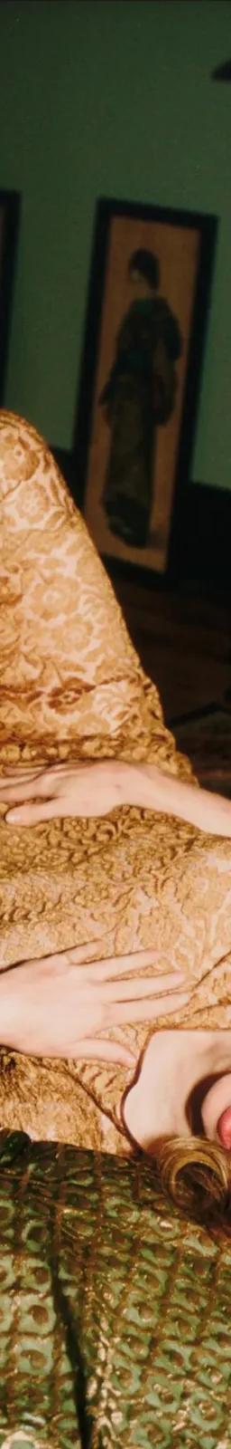 Someone in a gold lace dress lies on patterned green cushions in a dimly lit room, from Nan Goldin's photographic archive