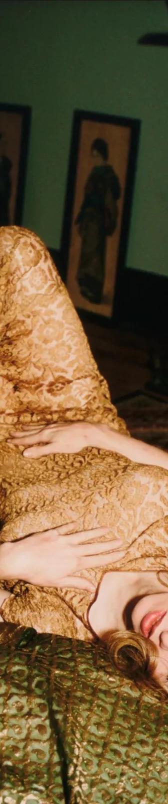 Someone in a gold lace dress lies on patterned green cushions in a dimly lit room, from Nan Goldin's photographic archive