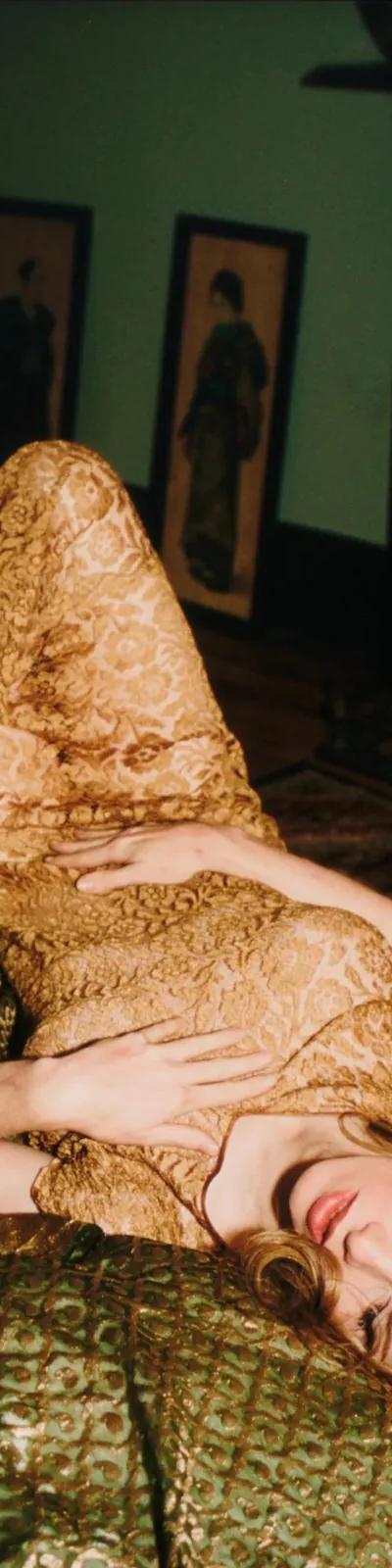 Someone in a gold lace dress lies on patterned green cushions in a dimly lit room, from Nan Goldin's photographic archive