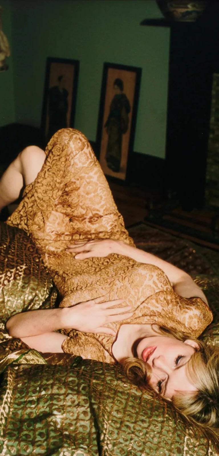 Someone in a gold lace dress lies on patterned green cushions in a dimly lit room, from Nan Goldin's photographic archive