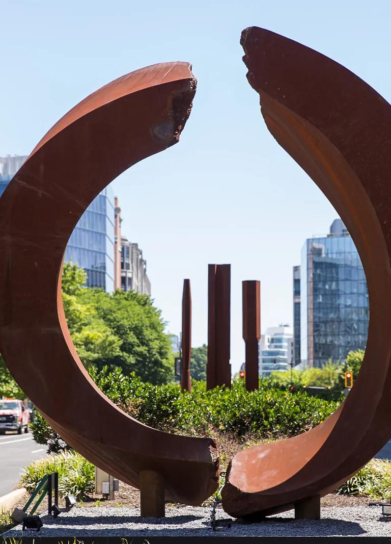 A circular, rust-colored sculpture and three rust-colored columns in the median of a city street.