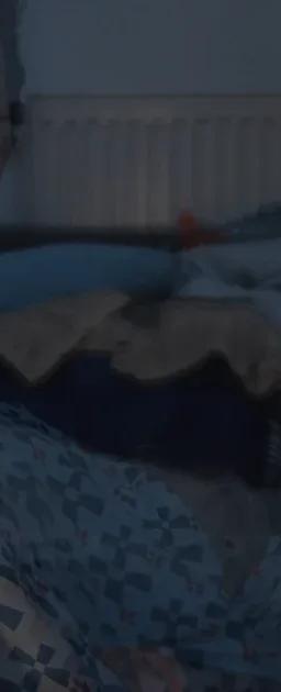 A translucent film still of an older person wrapped in a blue, white and red patterned blanket, in a dimly lit room with a radiator and piled blankets