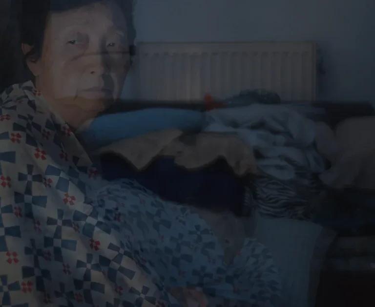 A translucent film still of an older person wrapped in a blue, white and red patterned blanket, in a dimly lit room with a radiator and piled blankets