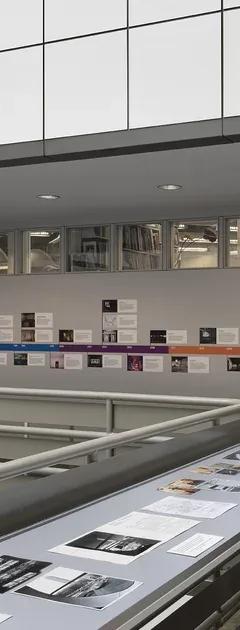 A large vitrine encasing black and white photographs in the middle of an open atrium, with a colourful illustrated timeline adhered to the wall behind