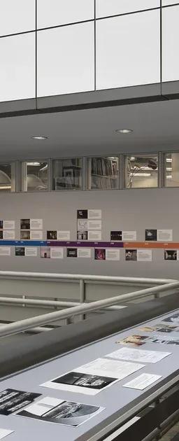 A large vitrine encasing black and white photographs in the middle of an open atrium, with a colourful illustrated timeline adhered to the wall behind