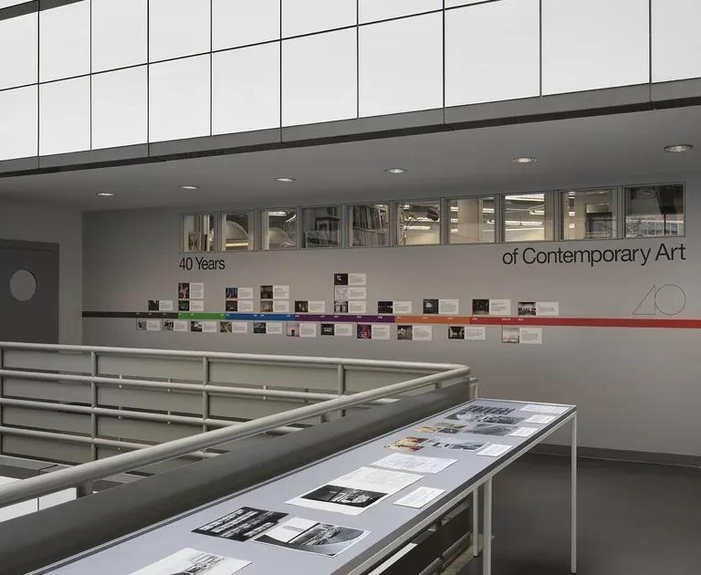 A large vitrine encasing black and white photographs in the middle of an open atrium, with a colourful illustrated timeline adhered to the wall behind
