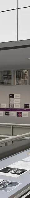 A large vitrine encasing black and white photographs in the middle of an open atrium, with a colourful illustrated timeline adhered to the wall behind