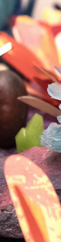 A close-up of a small, light blue crystal formation displayed on a rock, surrounded by colorful, paper-like flower shapes and decorations on a purple textured surface.