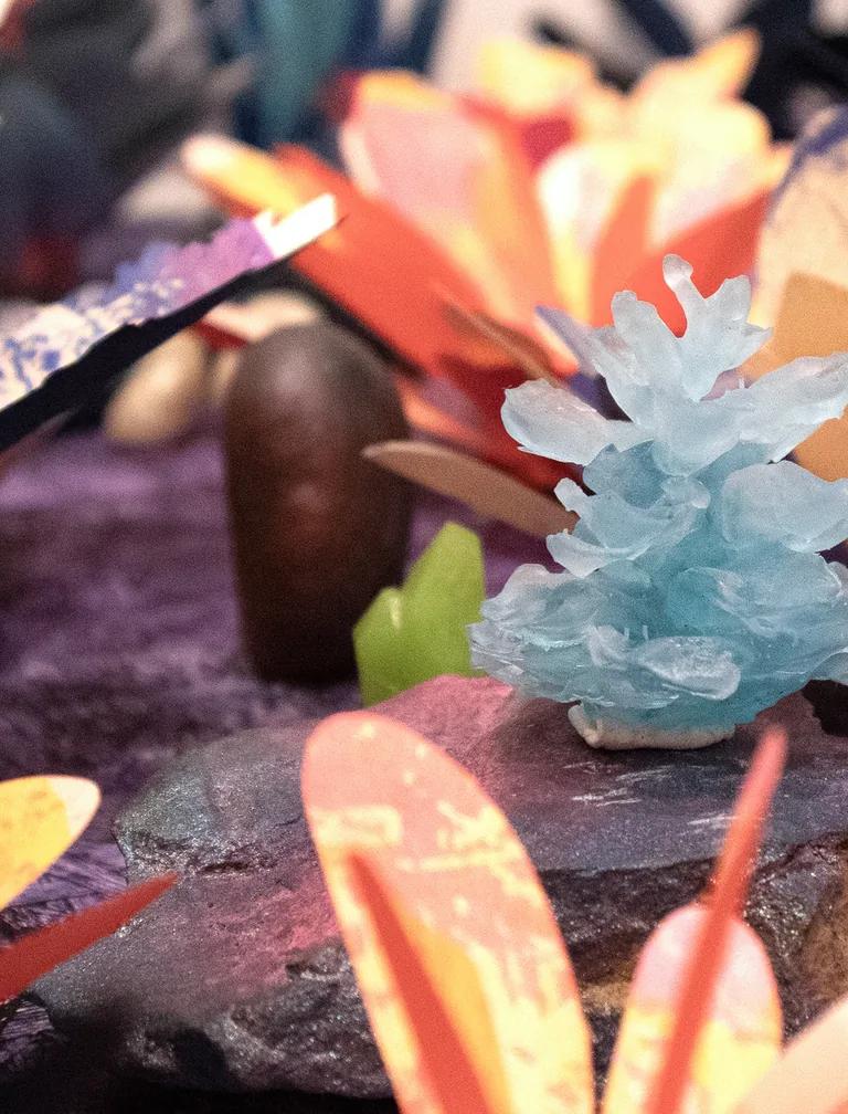 A close-up of a small, light blue crystal formation displayed on a rock, surrounded by colorful, paper-like flower shapes and decorations on a purple textured surface.