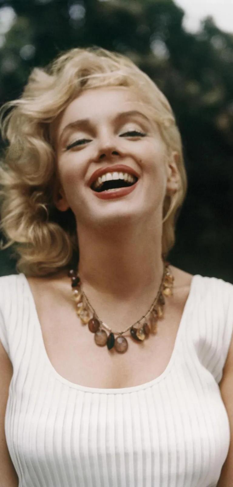 Colour photograph of Marilyn Monroe from the waist up, short blonde hair blowing in the wind, parted red lips, wearing a white ribbed vest and beaded necklace.